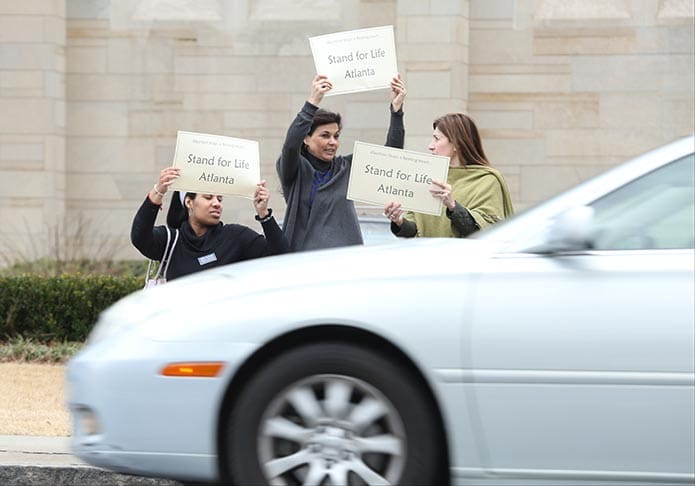 Cars whiz by as (l-r) Tiffany Bolado, Connie Alvarez and Maria da Silva of the Cathedral of Christ the King hold their signs on the east side of Peachtree Road during Stand for Life. Photo By Michael Alexander