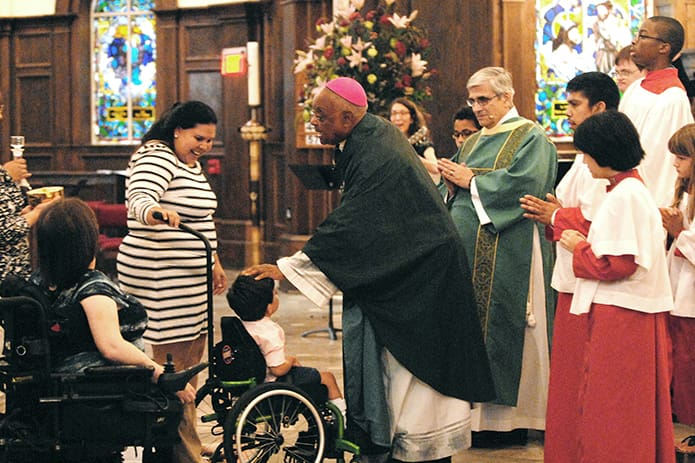 Archbishop Wilton D. Gregory blesses Lars Olivieri, 4, who has cerebral palsy and hydrocephalus, and his mother, Larissa Olivieri, as they lead the presentation of the gifts at the biannual Faith and Sharing Mass. In his homily, the archbishop said that the members of the disabilities community “remind us that our table, the Eucharist, the altar, will never be complete if we don’t invite you to come there.” PHOTO BY LAURA MOON.