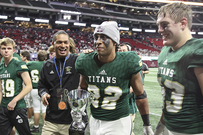 As Blessed Trinity running back Steele Chambers (#22) walks off the field and back to the locker room with offensive center Jack Filipowicz, right, carrying the championship trophy, he gets a congratulatory pat on the shoulder from Cornell Dore, left, a member of Blessed Trinityâs athletic association. Photo By Michael Alexander