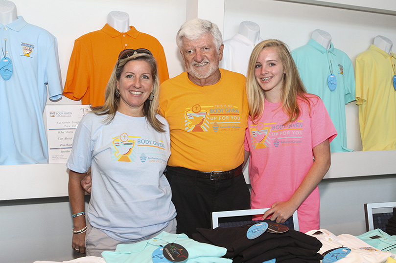 Tom Carroll, center, his daughter Jennifer Rollauer, left, and his 13-year-old granddaughter Kate represent three generations of T-shirt vendors at the Eucharistic Congress. Carrollâs company, TC Marketing, has been selling shirts at the annual event for over a decade. Recently diagnosed with Lymphoma, Carroll didnât want to miss this yearâs event as he prepares for cancer treatment.  Photo By Michael Alexander