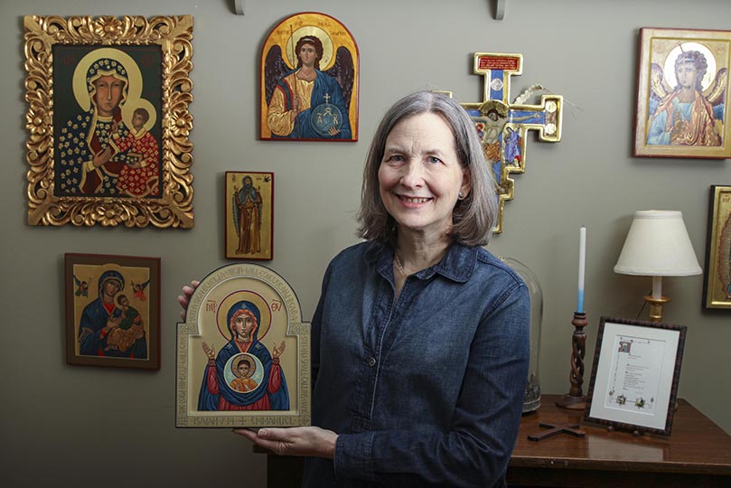 Icon painter and Holy Spirit Church parishioner Nancy Ewing holds one of her paintings, Our Lady of the Sign. The pieces hanging on the wall to her right, our left, were all painted by Ewing. They include (clockwise, from bottom left) Our Lady of Perpetual Help, Our Lady of Czestochowa, St. Michael the Archangel and Elijah and the Raven. Photo By Michael Alexander