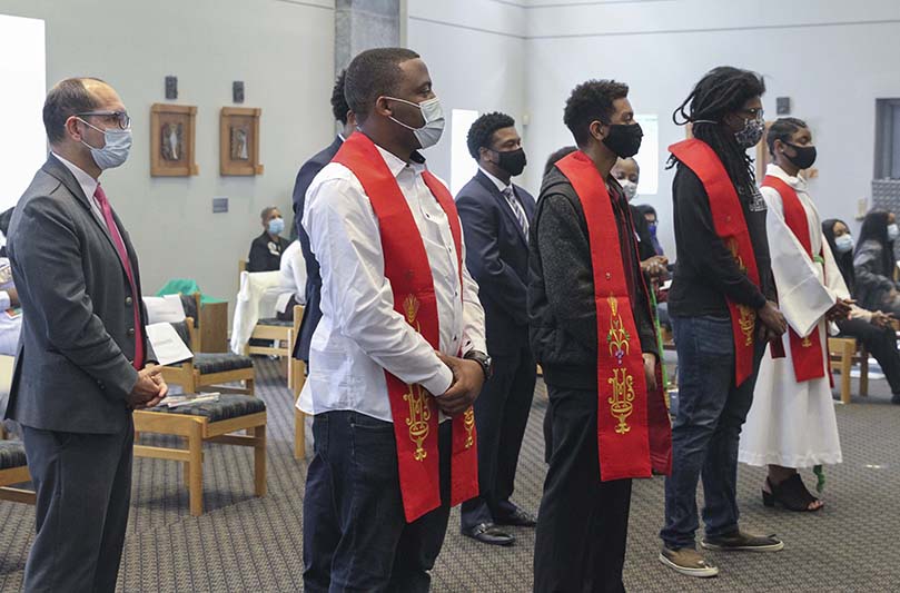 Wearing red confirmation stoles, the newly confirmed students stand before the altar at Lyke House Oct. 18. They included (front row, l-r) Moses Obenofunde, Christian Boothman, Douglas Geiger and Margaret Quartey. Obenofunde, Boothman and Geiger are Georgia State University students. Quartey, the niece of Lyke House chaplain and director, Father Urey Mark, is an Our Lady of Mercy High student and a frequent altar server at the Catholic Center. Obenofunde, Boothman and Geiger were also baptized into the faith. Photo By Michael Alexander