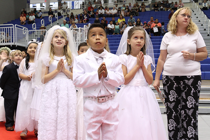 (Foreground center, l-r) Eight-year-olds Anna Claire Beverly and Michael Gonzalez and 7-year-old Emma McWhorter watch themselves overhead on the jumbo screen at the University of West Georgia Coliseum, Carrollton, as they prepare to lead the opening procession of first communicants to their seats. They made their first holy Communion with 73 other youngsters from Our Lady of Perpetual Help Church on May 1, the sixth Sunday of Easter. Photo by Michael Alexander