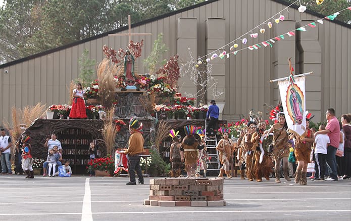 Danza De Apaches dancers of Lawrenceville, right, perform to a drummer in the back parking lot at Our Lady of the Americas Mission, Lilburn, as spectators watch and others organize the flowers atop the huge shrine honoring Our Lady of Guadalupe. Photo By Michael Alexander