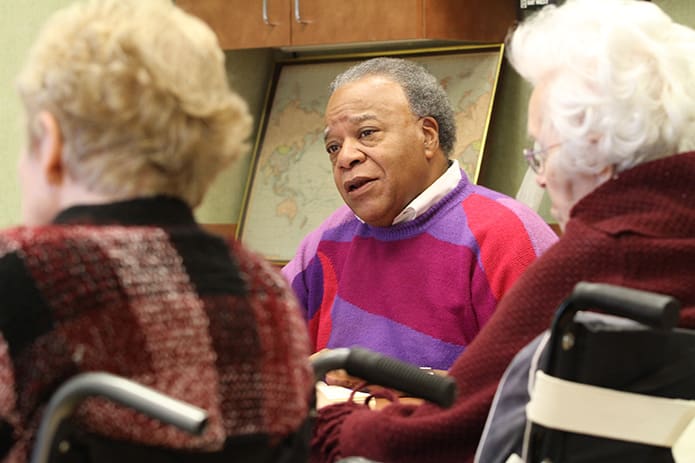 Sitting in their wheelchairs, Mary Warren, left, and Virginia Stearns listen to David Waters, center, provide a brief synopsis of the Gospel from Luke. Waters reads the dayâs Gospel before they pray the rosary. He and his wife Mary began participating in 2006 when his mother was a resident at St. George Villageâs Wellington Court. In 2007 Waters became the unofficial facilitator for the ministry, which he continues even though his mother died on All Saints Day in 2012. Photo By Michael Alexander