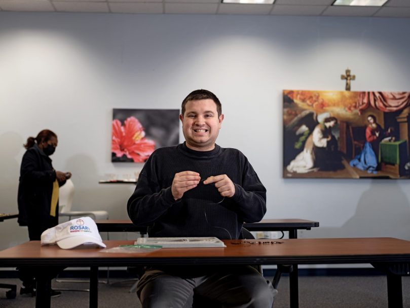 Michael Feliu pauses work to share a smile while making a rosary at the Rosary Makers of America office. Photo by Johnathon Kelso