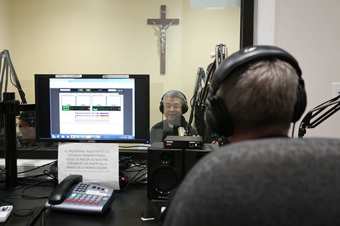 From the other side of the glass, Father Luis Guillermo CÃ³rdoba, background, administrator of Our Lady of the Americas Mission, converses with Bishop Luis R. Zarama, foreground, during programming of the Spanish Catholic radio program, âNuestra Fe.â  Photo By Michael Alexander