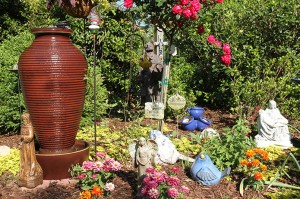 In one part of the Nelson garden among the flowers and shrubs are (foreground, l-r) a statue of St. Francis, an angel, and a statue of the Pieta. In the background is another statue of St. Francis, some crosses and a sign that says, 